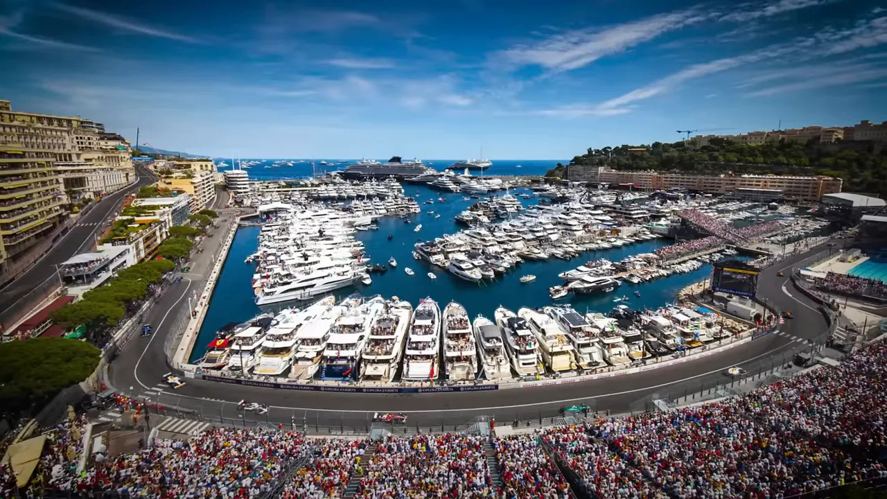 Aerial view of Monaco harbour filled with yachts alongside the Monaco Grand Prix street circuit and packed grandstands under a blue sky.
