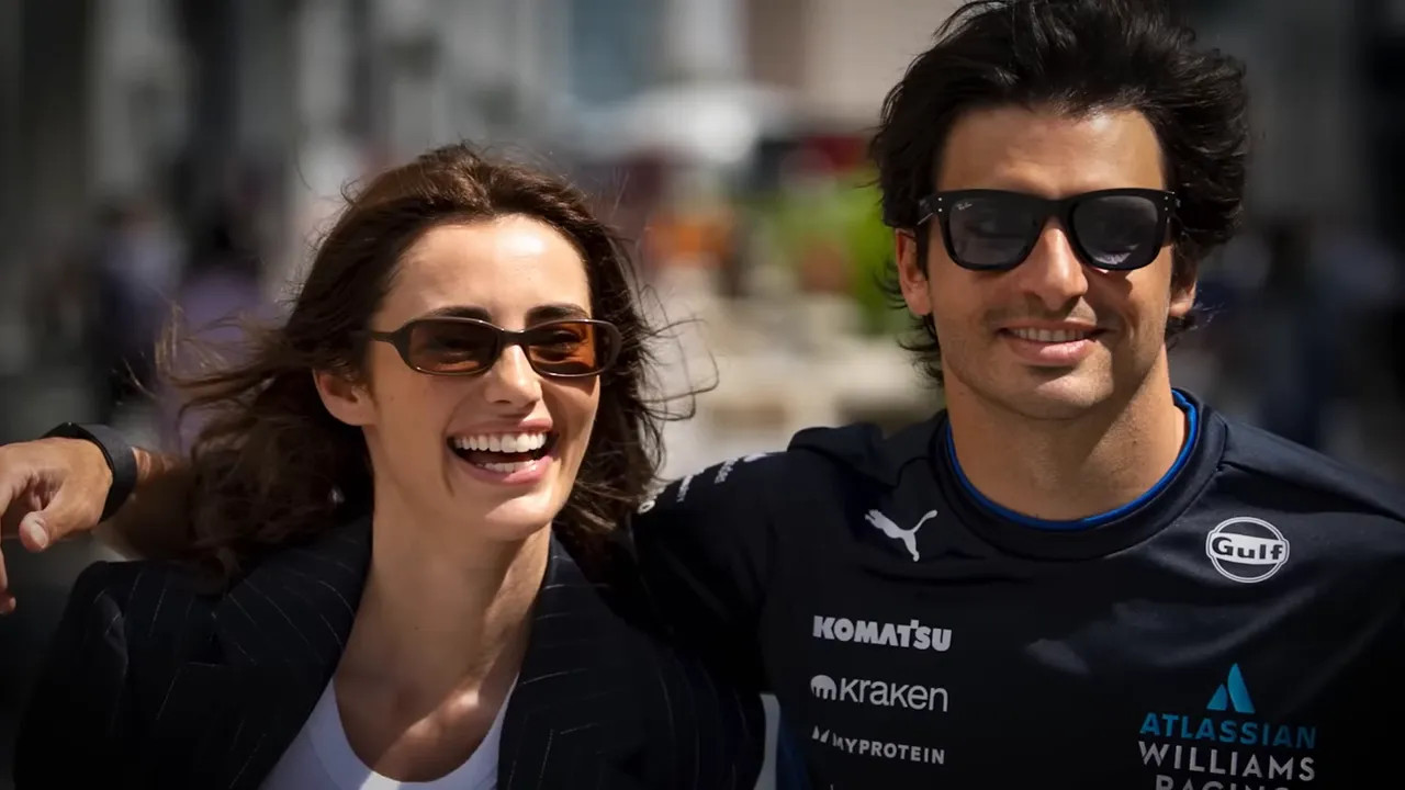 Driver and his partner smiling and walking arm-in-arm through the paddock, both wearing sunglasses