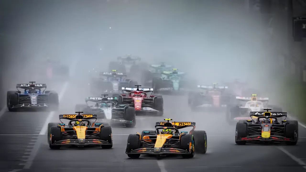 Formula 1 cars racing through heavy spray at the start of a wet Grand Prix with multiple cars visible in a tight pack