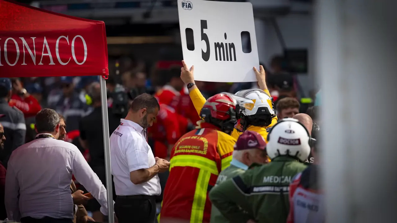 Track official in yellow and red overalls holding a large 5 min sign in pit lane