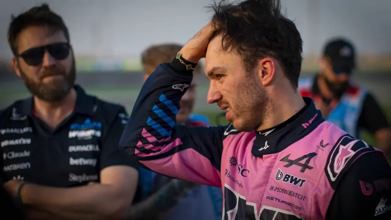 Driver in a pink race suit rubbing his head and looking concerned while team members stand behind