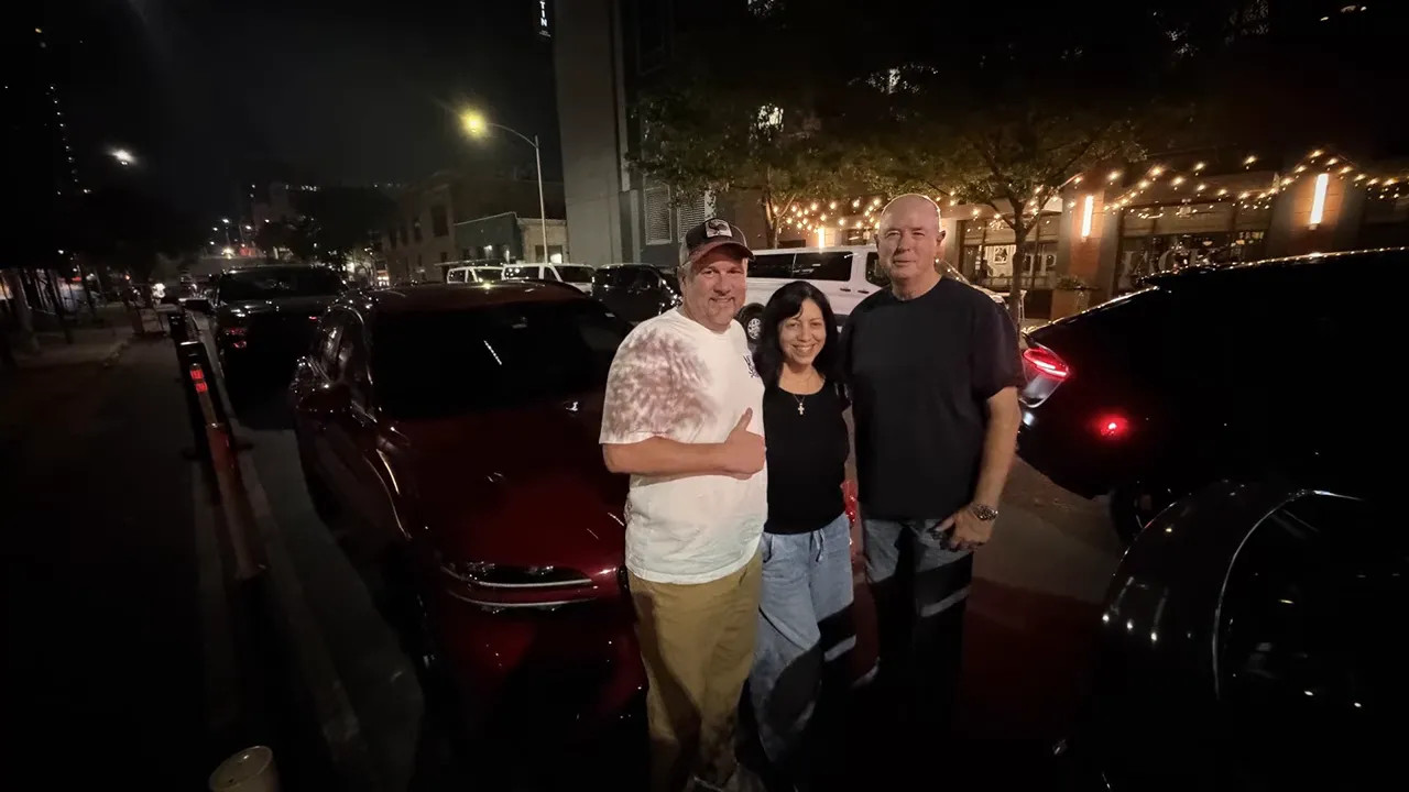 3 people standing beside a red car in a car park at night