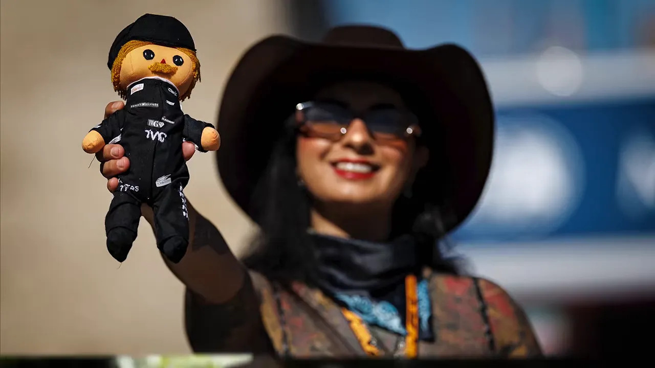 A woman wearing a cowboy hat and sunglasses holds 2 handmade racing driver dolls