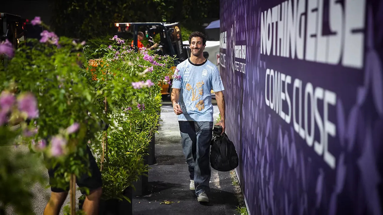 Daniel Ricciardo carrying a black bag along a narrow paddock path bordered by flowers