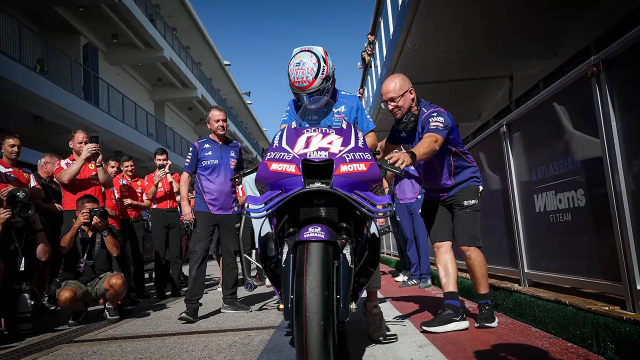 Franco Colapinto seated on a purple MotoGP motorcycle while a crew member pushes from behind