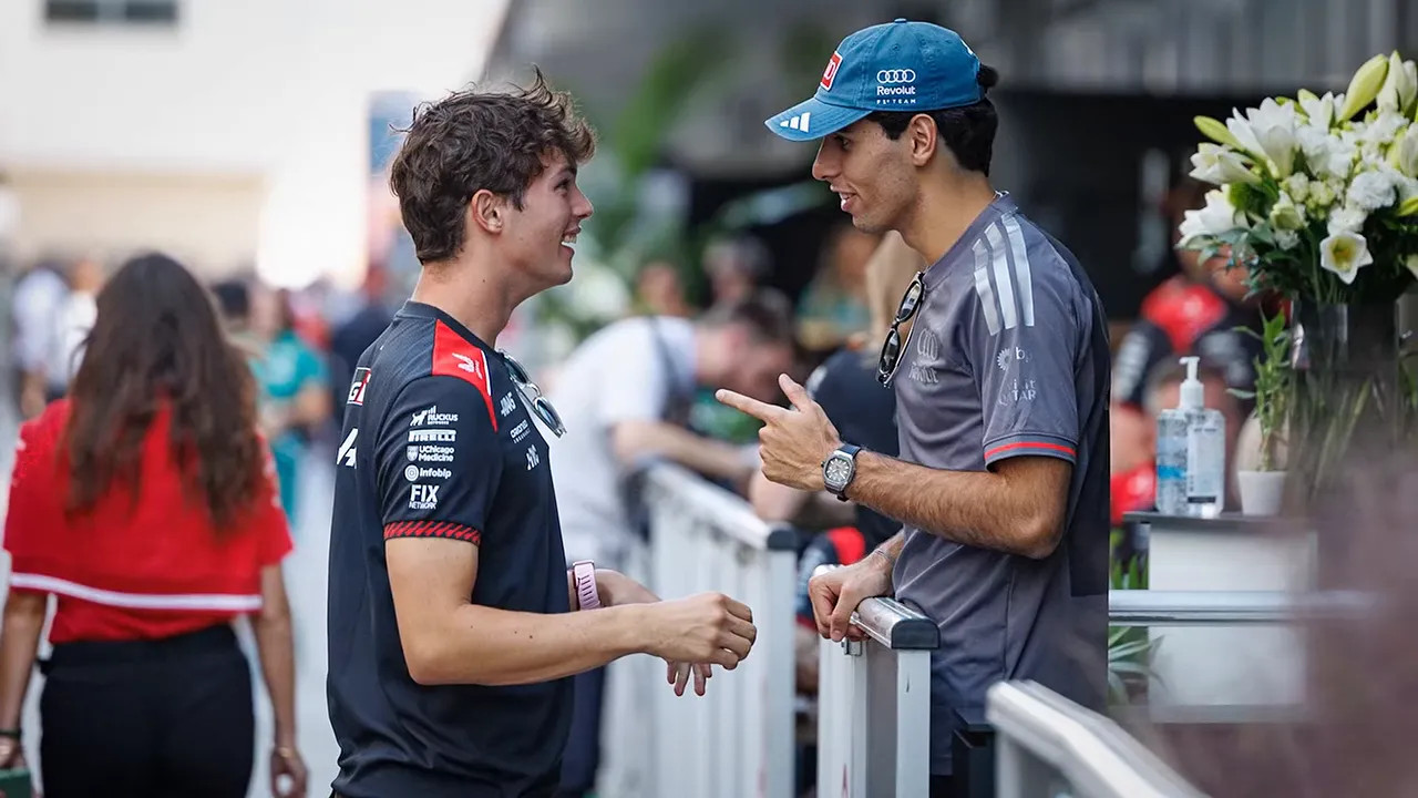 Ollie Bearman and Gabriel Bortoleto laughing together beside a paddock railing