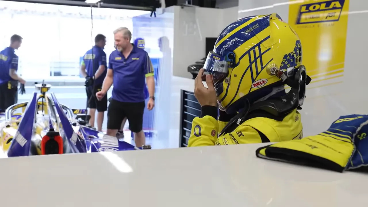 Driver in a yellow racing suit adjusting their visor while a blue-and-yellow single-seater and crew are visible in the garage.