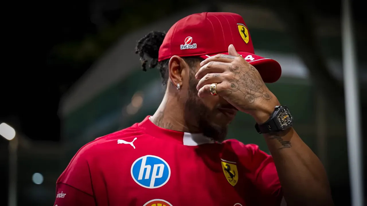 Ferrari driver in team apparel and cap covering his face with his hand, showing clear frustration.