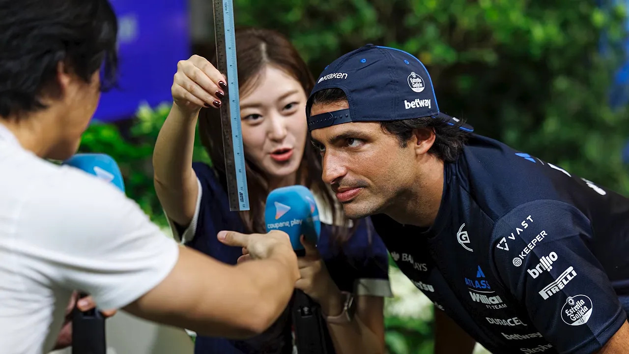Carlos Sainz holding a ruler during a reaction test surrounded by microphones
