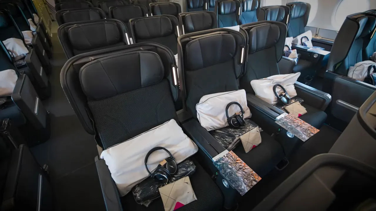 Aircraft mid‑cabin seats arranged in a 2-4-2/2-3-2 style with pillows, headphones and amenity packs on each seat.