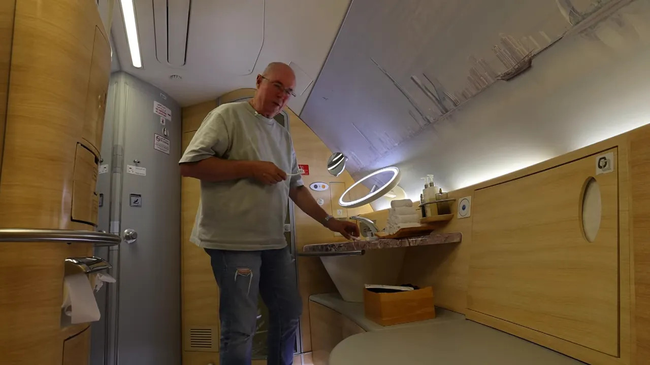 First‑class aircraft lavatory showing a sink, illuminated mirror, stacked towels and toiletry dispensers with a person at the basin.