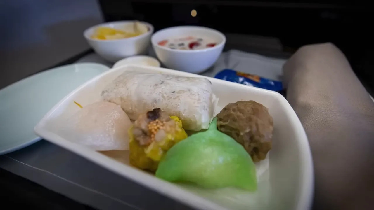 tray of in-flight dim sum and sides including a green dumpling, siu mai and bowls of fruit and cereal on a tray table