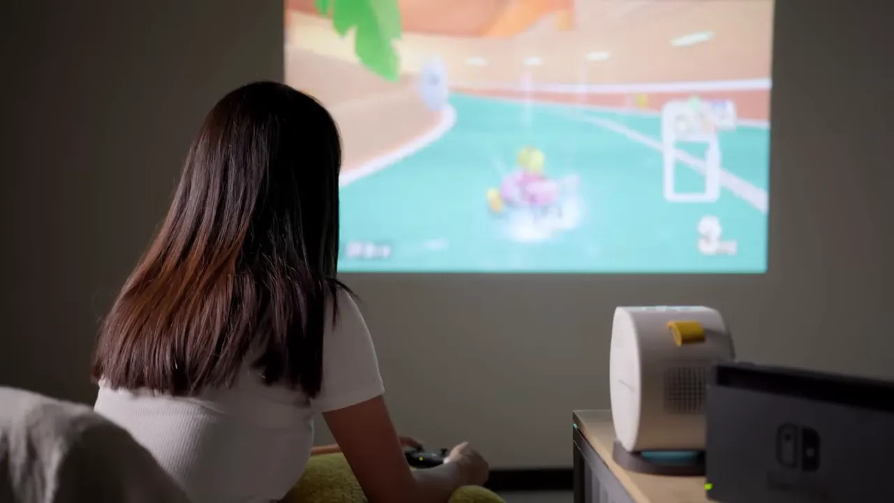 Person seated playing a racing game projected on a wall, viewed from behind