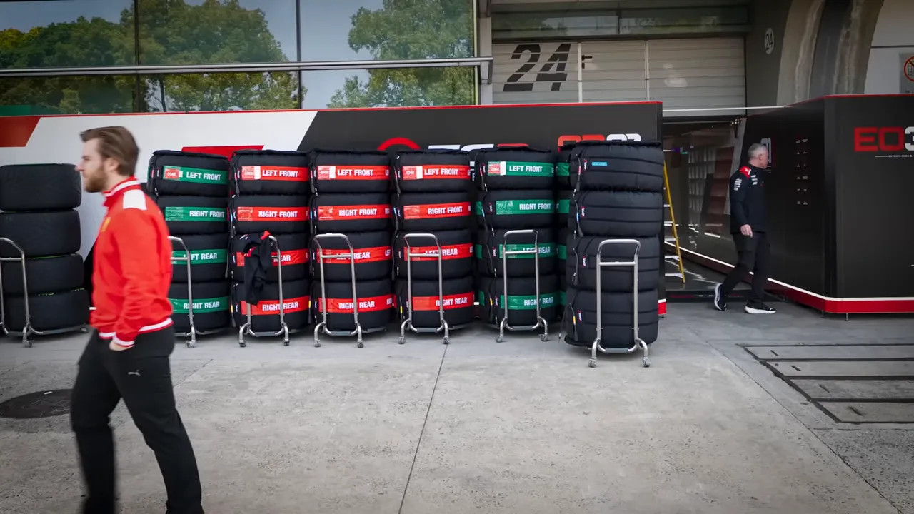 Stacks of tyre sets on trolleys outside a team garage in the paddock, with team staff walking by.