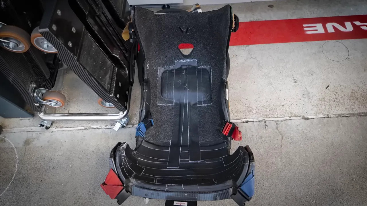 Top-down view of a Formula 1 seat shell on a garage floor with taped ballast areas and harness anchors