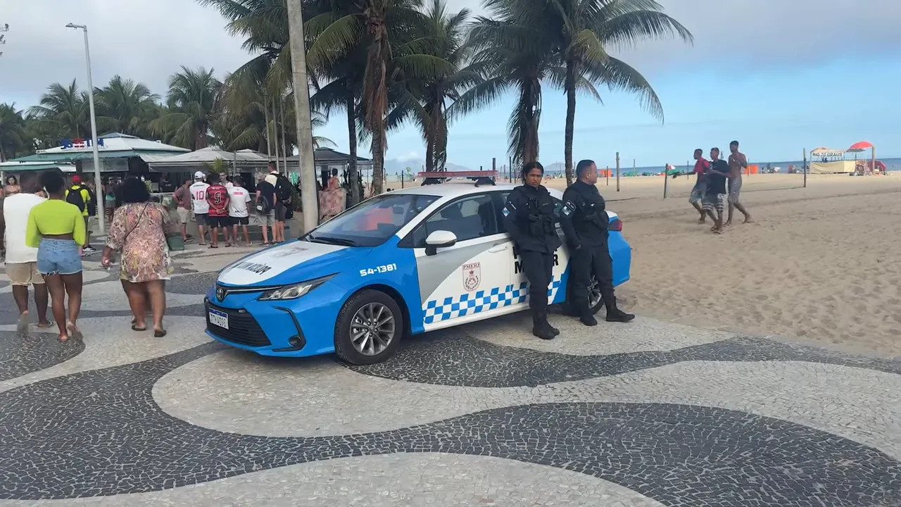 Two police officers standing by a blue-and-white patrol car on a beach promenade