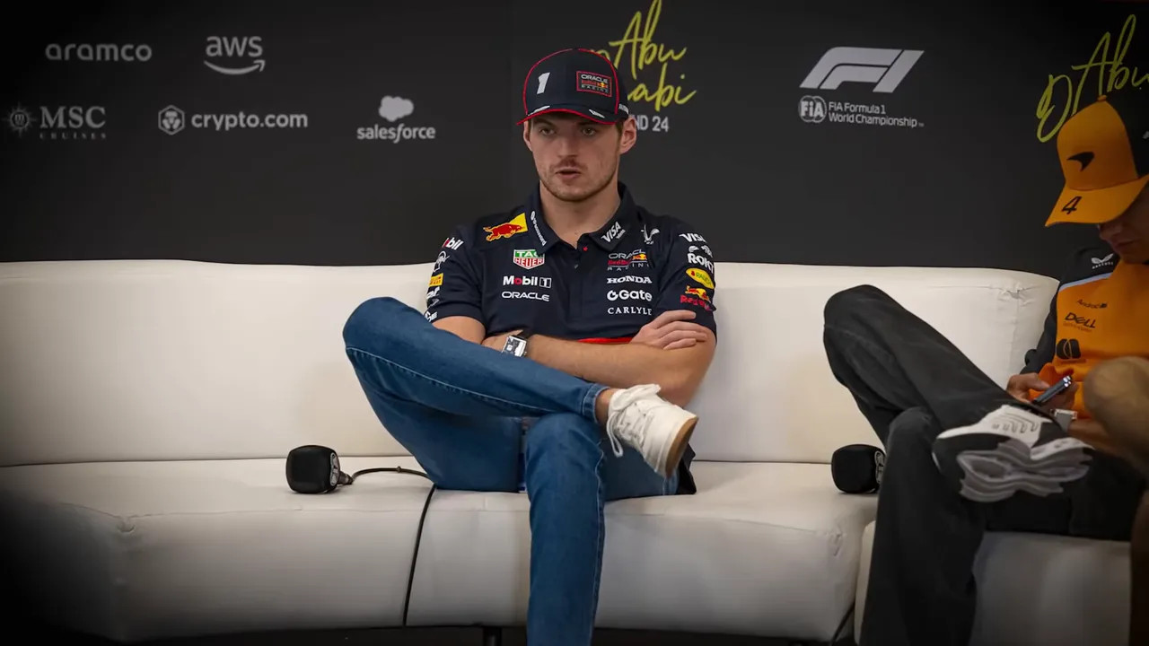 Formula 1 driver seated on a white couch in a press area with legs crossed and arms folded, showing a composed posture.