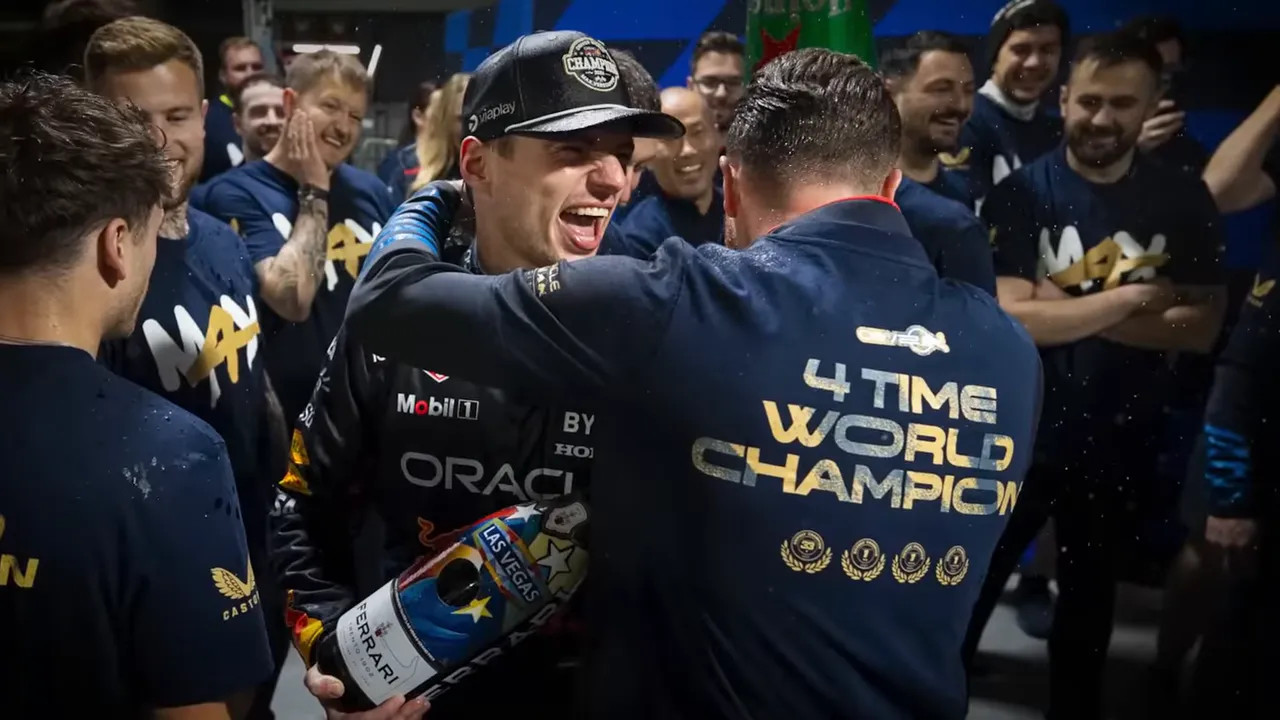 Team members celebrating in a garage while one person wears a jacket reading '4 TIME WORLD CHAMPION'