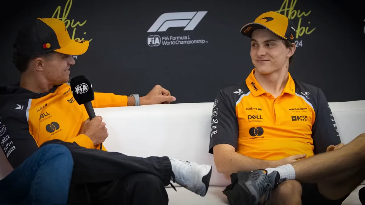 Two McLaren drivers seated on a couch during a press conference, one holding an FIA microphone and both in team kit.