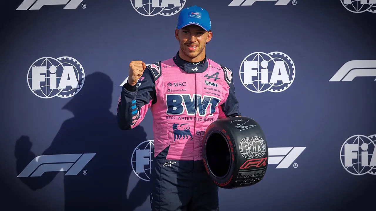 Pierre Gasly in pink Alpine race suit holds a helmet and raises a clenched fist
