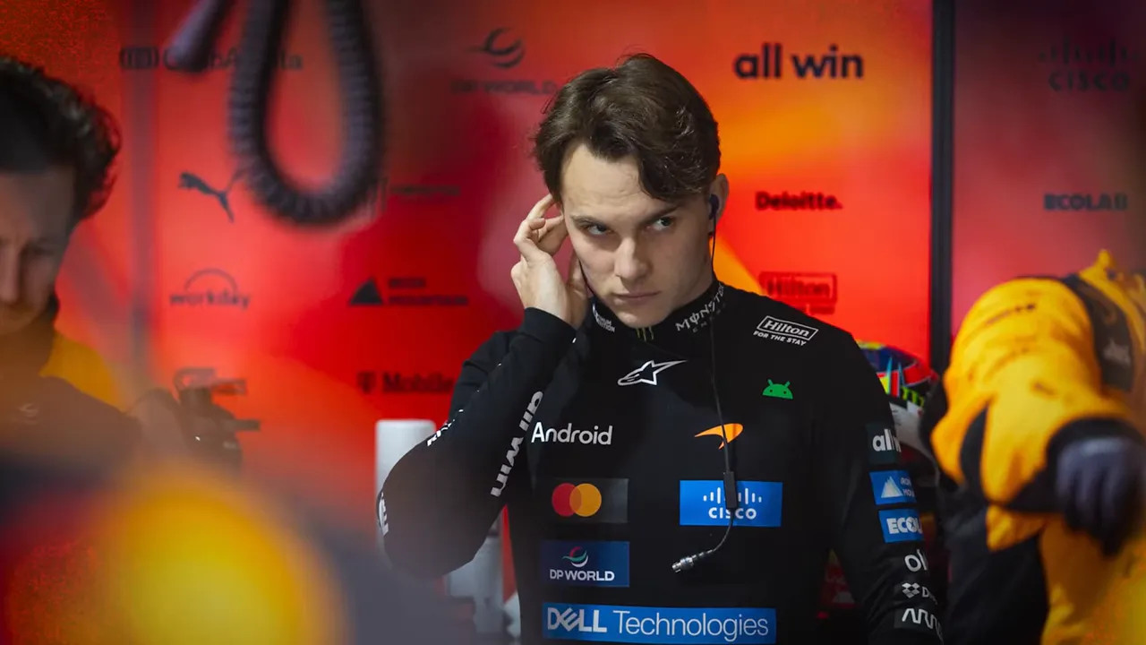 Focused McLaren driver in the team garage adjusting an earpiece, framed by orange team colours.