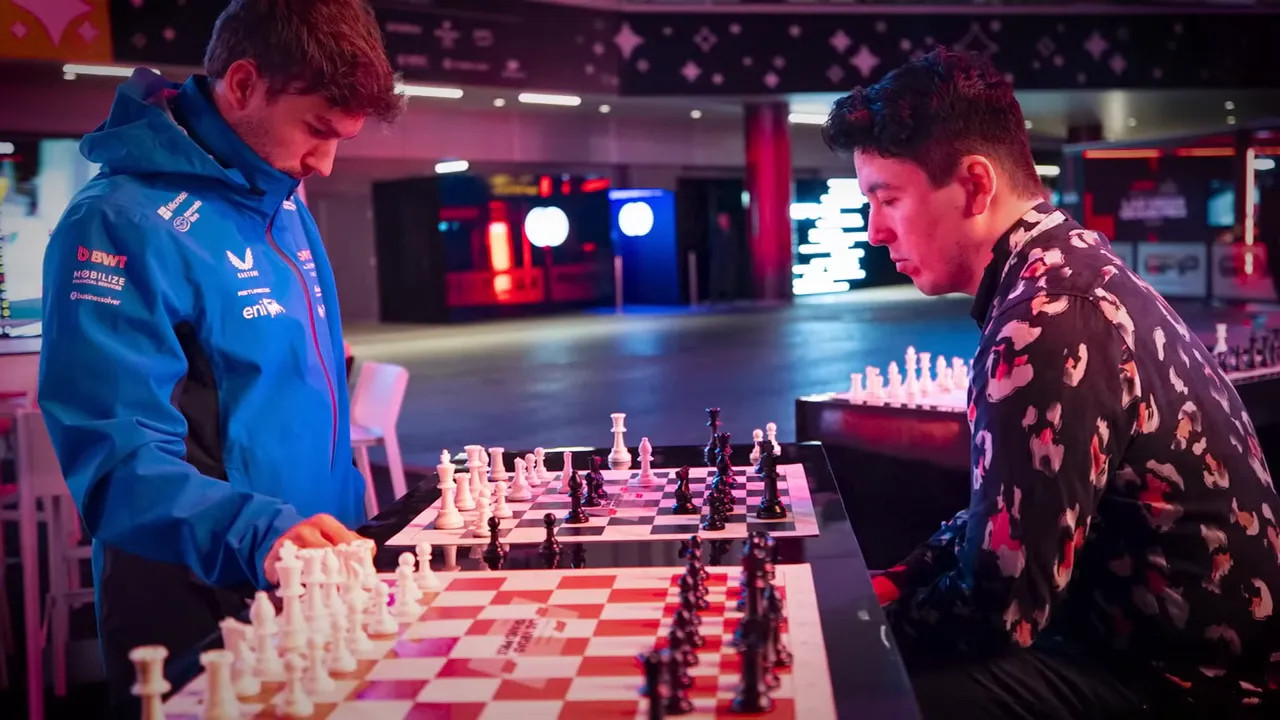Two players competing across several chess boards in the paddock with the illuminated circuit concourse behind them.