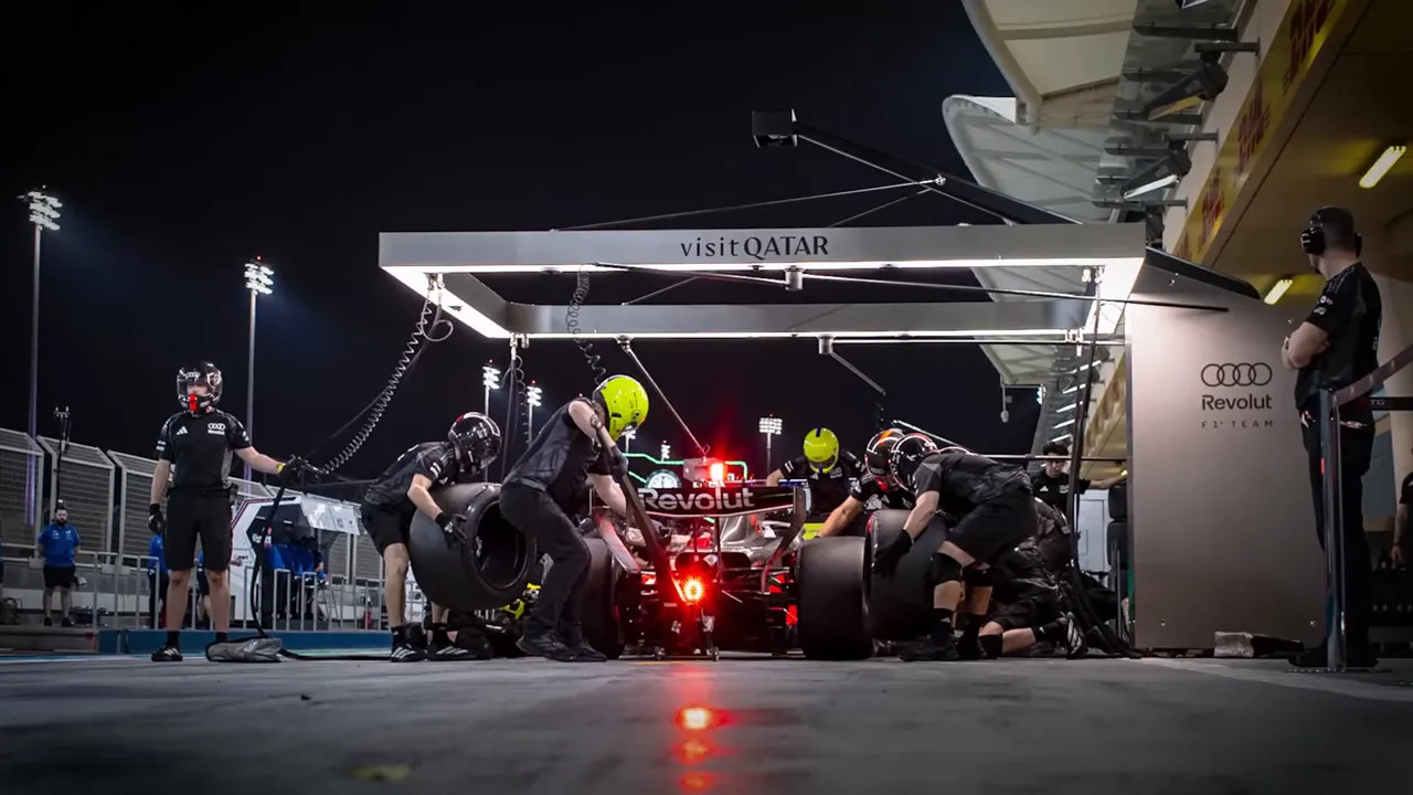 Rear view of an Audi pit stop under an illuminated 'visit QATAR' gantry with crew and car at night.