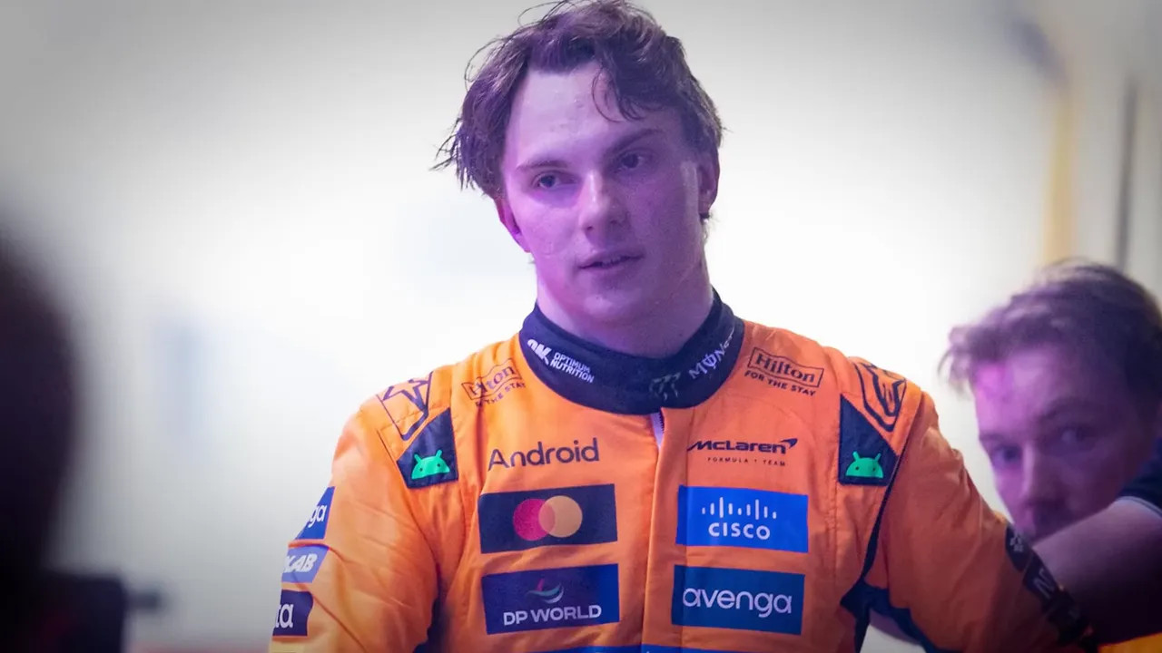 Head-and-shoulders of a McLaren driver in an orange race suit looking frustrated and reflective in the garage