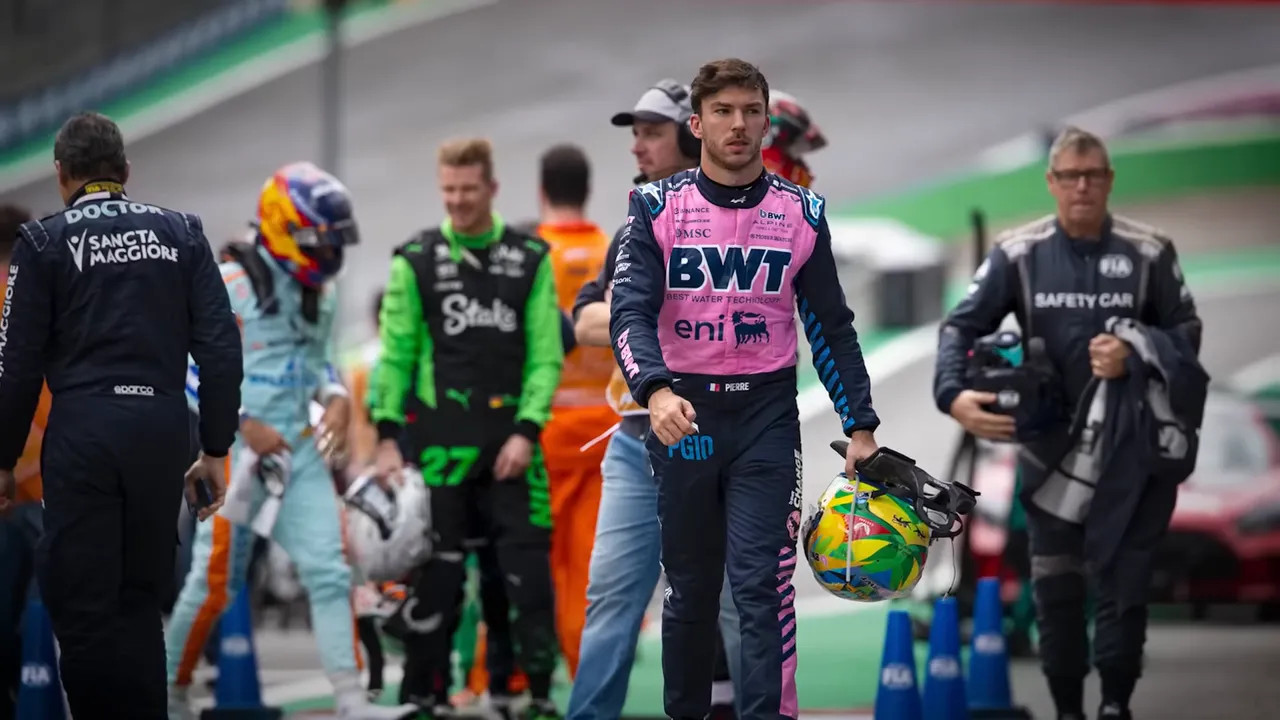 Driver walking in the pit lane holding a helmet with team members and officials in the background