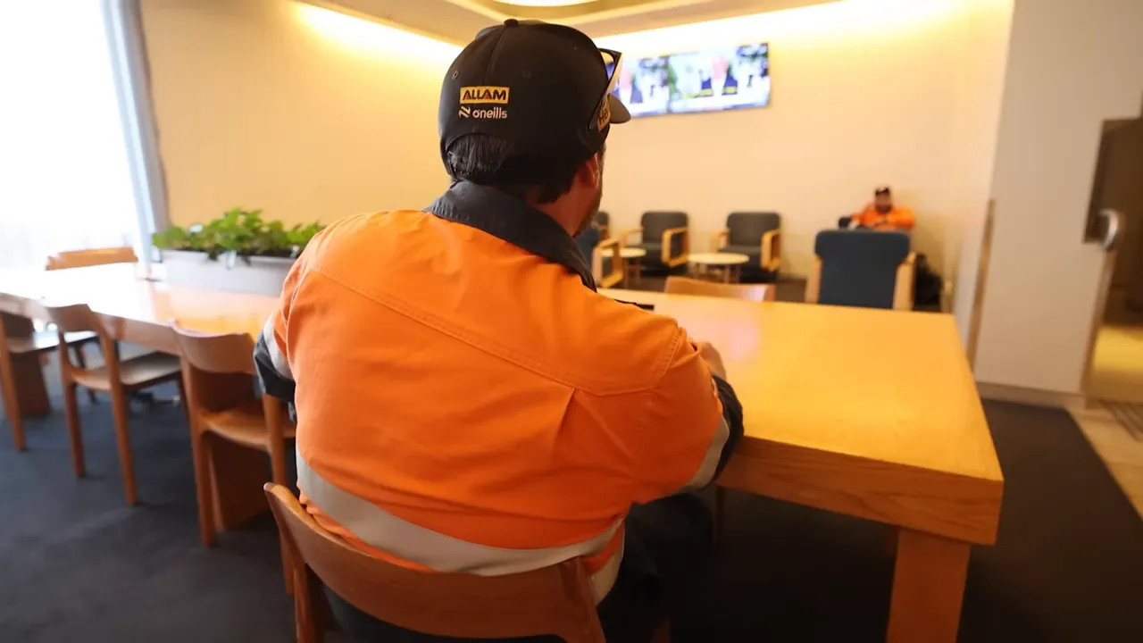 Back view of a passenger wearing an orange high‑visibility jacket and cap seated at a wooden table in an airport lounge