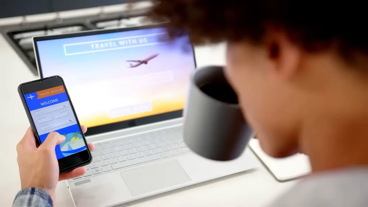 Person booking travel on a smartphone with a laptop showing an airplane image