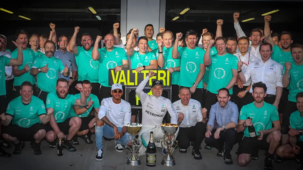 Driver kneeling with trophies and surrounded by the celebrating team in the garage after a race victory.