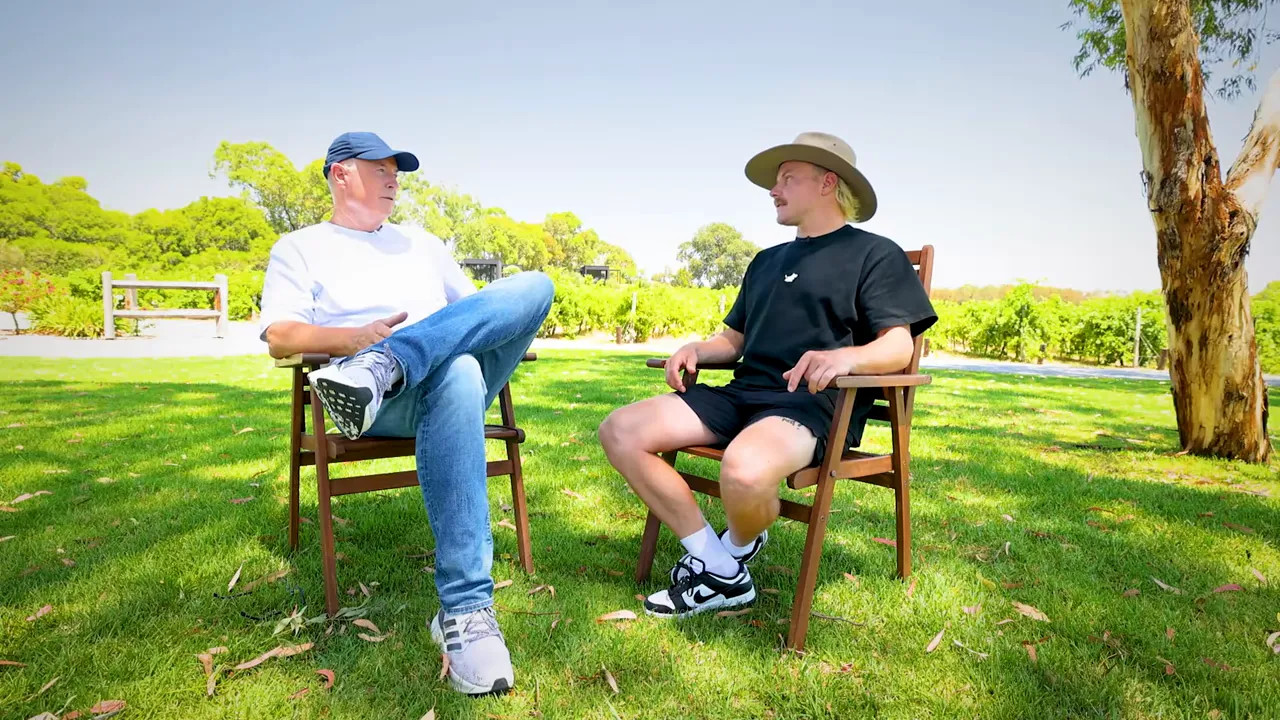 Two people seated on the grass in wooden chairs in front of vineyard rows engaged in conversation
