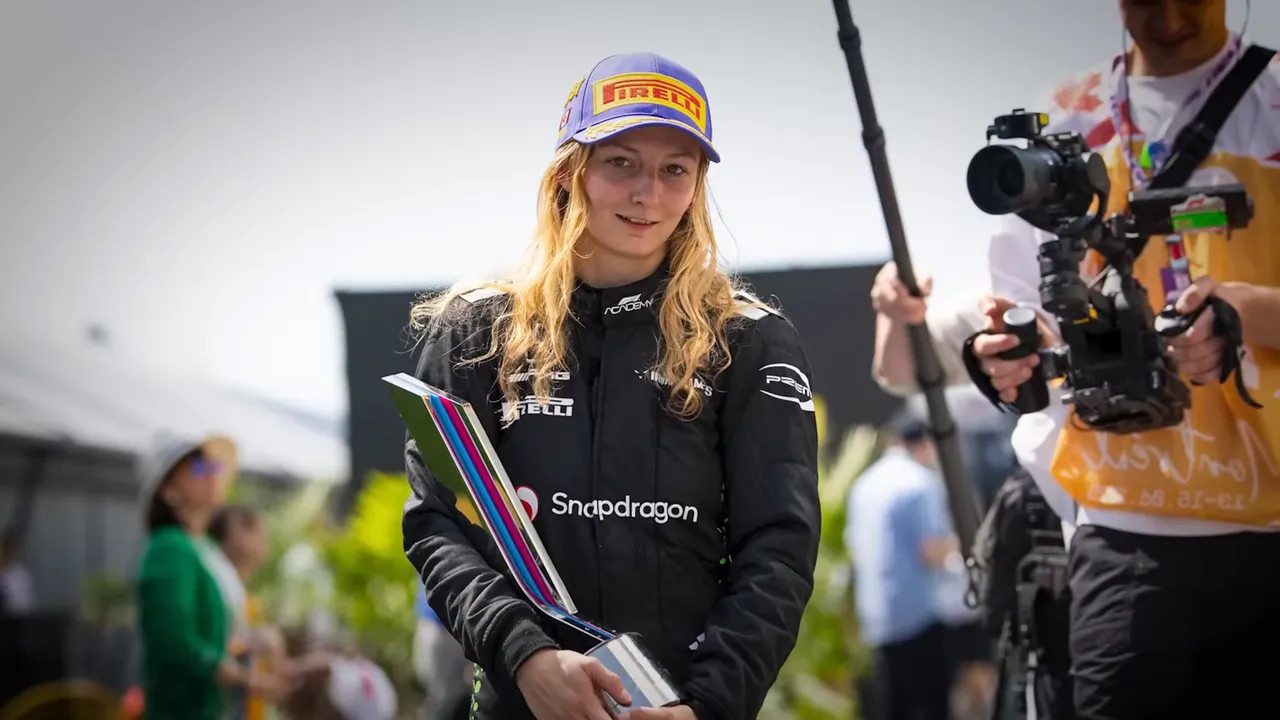 F1 Academy driver in racing overalls holding a trophy with media and onlookers in the background.