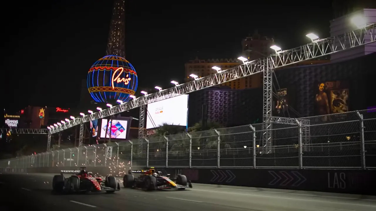 Two Formula cars racing on the Las Vegas Strip at night beneath an overhead truss with sparks flying