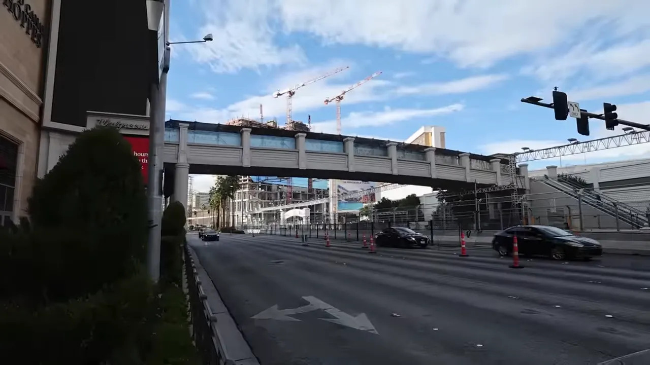 Wide shot of a pedestrian overpass crossing the Las Vegas Strip with temporary spectator fencing and construction cranes in the background.