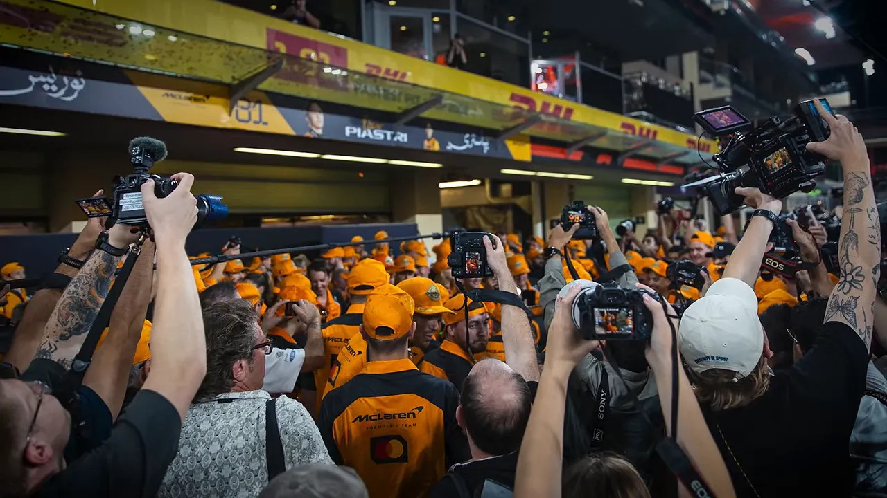 Photographers raising cameras around a crowded McLaren celebration outside a garage