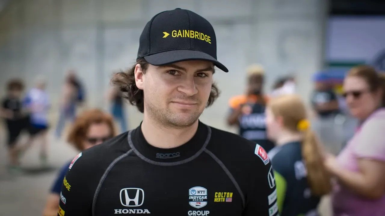 Driver wearing a Gainbridge cap and racing top standing in the paddock with fans blurred in the background.