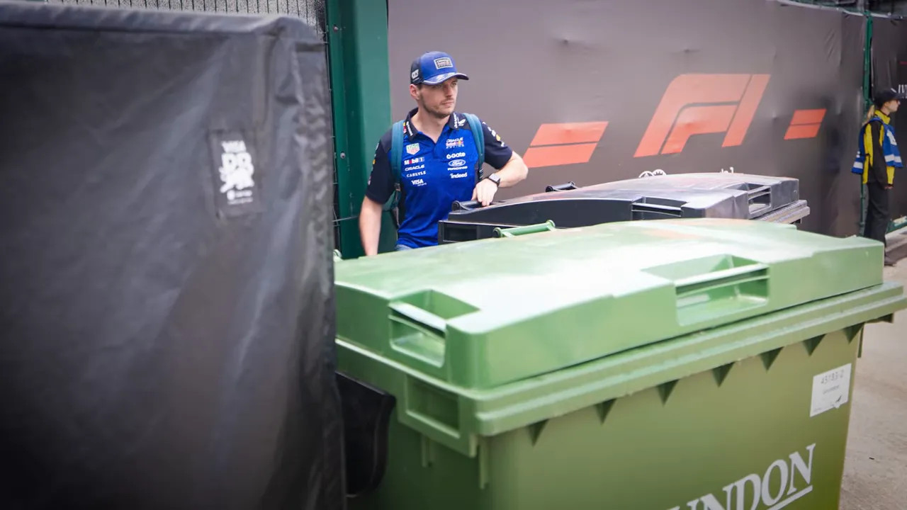 Max Verstappen standing near large green bins beside an F1 barrier wall