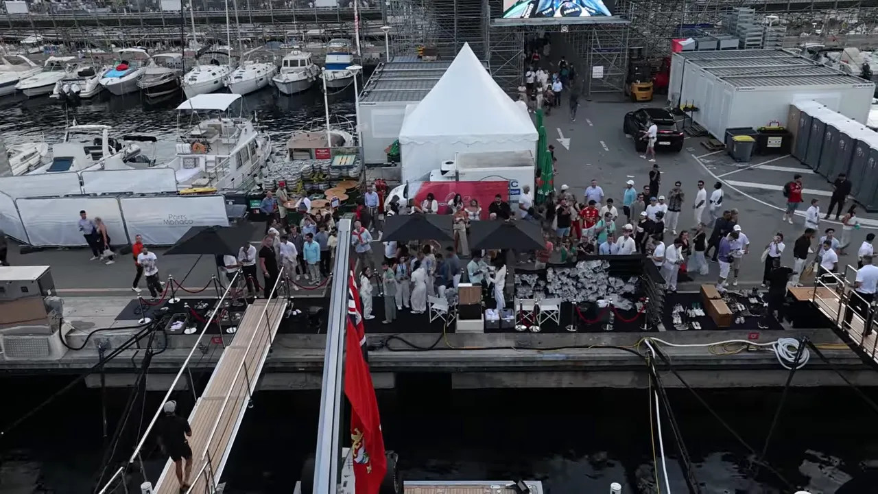 View down from the yacht toward a busy dock area with seated guests, a white tent, and harbour walkways
